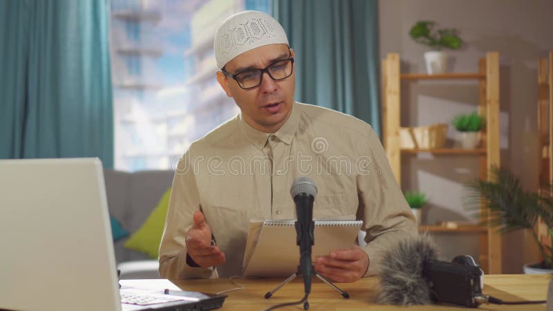 πορτραίτο μουσουλμάνος με καπέλο skulcap καταγράφει podcast ενώ κάθεται μπροστά από ένα μικρόφωνο στο σαλόνι