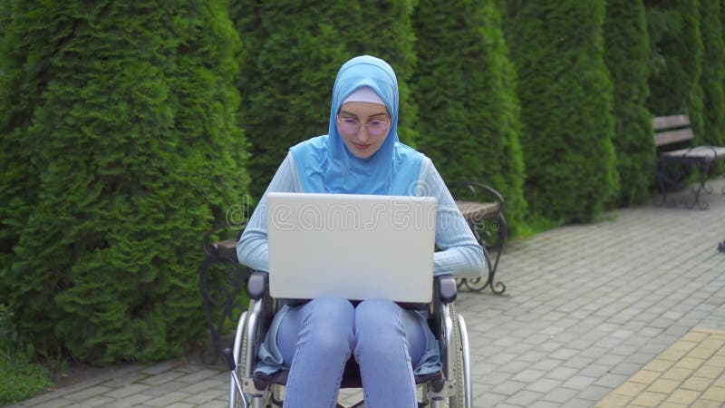 Πορτραίτο νεαρή μουσουλμάνα με ειδικές ανάγκες σε παραδοσιακό κασκόλ χρησιμοποιεί ένα φορητό υπολογιστή που κάθεται σε αναπηρική