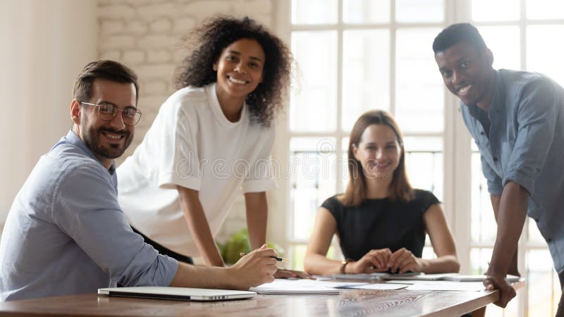 Head shot portrait young happy mixed race group of professionals sitting at table in modern office. Smiling multiracial leaders executives employees corporate people workers looking at camera. Head shot portrait young happy mixed race group of professionals sitting at table in modern office. Smiling multiracial leaders executives employees corporate people workers looking at camera.