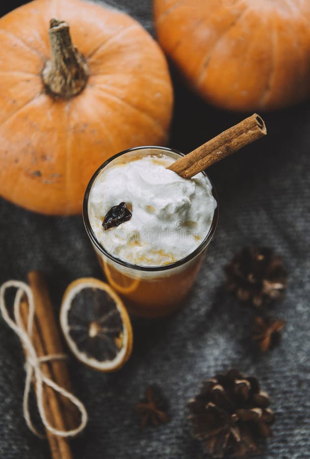 1 Cup fresh pumpkin juice with whipped cream and cinnamon stick, whole pumpkins, a slice of dried orange, cones on a knitted dark fabric, top view. 1 Cup fresh pumpkin juice with whipped cream and cinnamon stick, whole pumpkins, a slice of dried orange, cones on a knitted dark fabric, top view