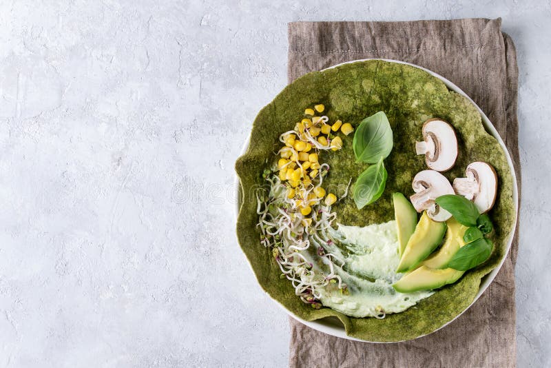 Green spinach matcha tortilla with vegan ingredients for filling. Sweet corn, avocado, green paprika, sprouts, mushrooms served in white plate over gray texture background and textile. Flat lay. Green spinach matcha tortilla with vegan ingredients for filling. Sweet corn, avocado, green paprika, sprouts, mushrooms served in white plate over gray texture background and textile. Flat lay
