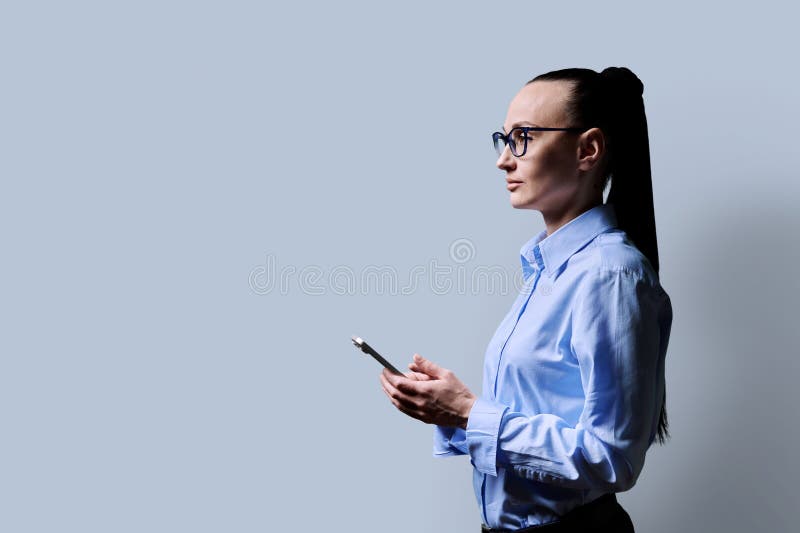 Profile view 30s serious woman using smartphone on grey background. Middle aged female in glasses looking forward. Technologies mobile apps applications internet work business leisure communication. Profile view 30s serious woman using smartphone on grey background. Middle aged female in glasses looking forward. Technologies mobile apps applications internet work business leisure communication