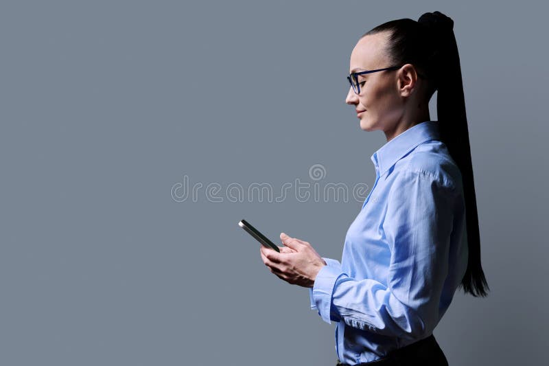 Profile view 30s smiling woman using smartphone on grey background. Middle aged female looking at phone in hands. Technologies mobile apps applications internet work business leisure communication. Profile view 30s smiling woman using smartphone on grey background. Middle aged female looking at phone in hands. Technologies mobile apps applications internet work business leisure communication