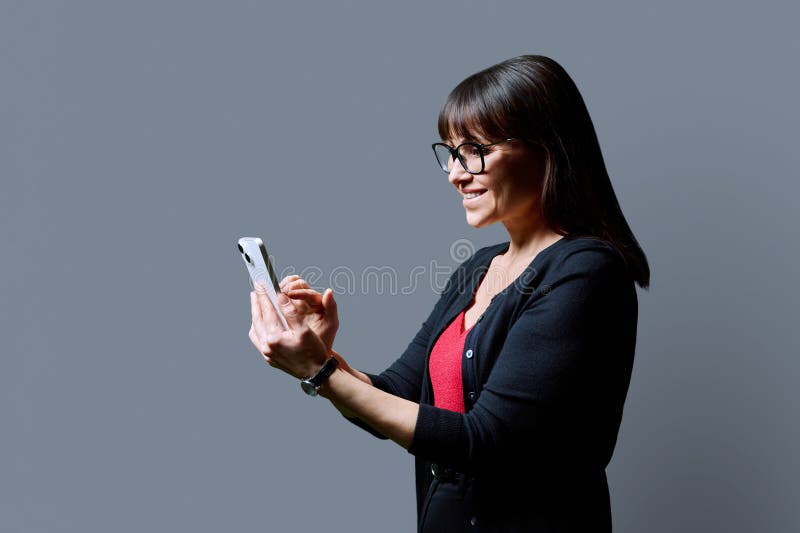 Profile view middle aged smiling woman using smartphone on grey background. Mature female looking at phone in hands. Technologies mobile apps applications internet work business leisure communication. Profile view middle aged smiling woman using smartphone on grey background. Mature female looking at phone in hands. Technologies mobile apps applications internet work business leisure communication