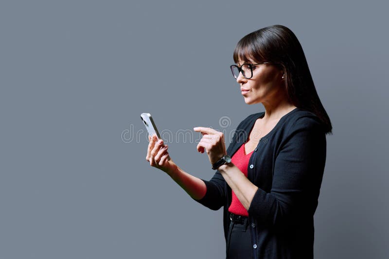 Profile view middle aged serious woman using smartphone on grey background. Mature female looking at phone in hands. Technologies mobile apps applications internet work business leisure communication. Profile view middle aged serious woman using smartphone on grey background. Mature female looking at phone in hands. Technologies mobile apps applications internet work business leisure communication