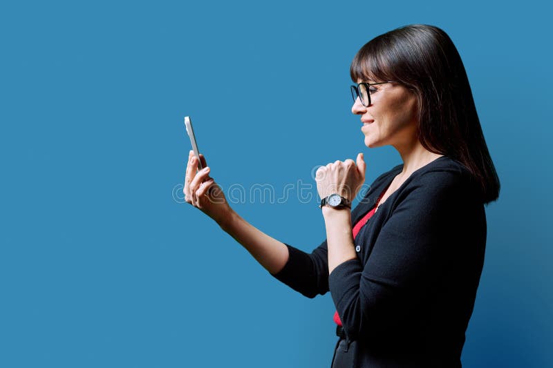 Profile view middle aged smiling woman using smartphone on blue background. Mature female looking at phone in hands. Technologies mobile apps applications internet work business leisure communication. Profile view middle aged smiling woman using smartphone on blue background. Mature female looking at phone in hands. Technologies mobile apps applications internet work business leisure communication