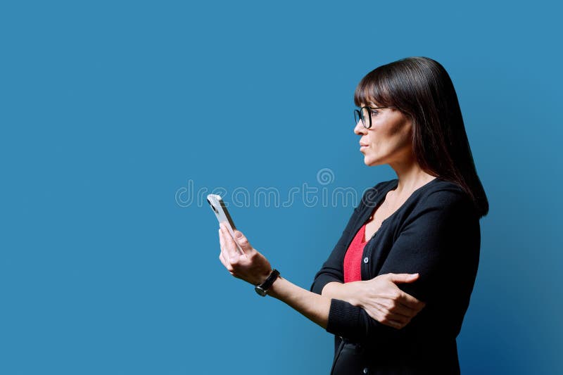 Profile view middle aged serious woman using smartphone on blue background. Mature female looking at phone in hands. Technologies mobile apps applications internet work business leisure communication. Profile view middle aged serious woman using smartphone on blue background. Mature female looking at phone in hands. Technologies mobile apps applications internet work business leisure communication