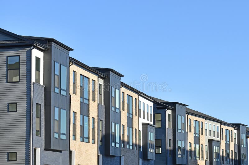 Landscaping and young evergreen trees accent these hip townhouses with painted brick, pale yellow unpainted brick, vinyl siding, painted wood, and garage doors. Landscaping and young evergreen trees accent these hip townhouses with painted brick, pale yellow unpainted brick, vinyl siding, painted wood, and garage doors.