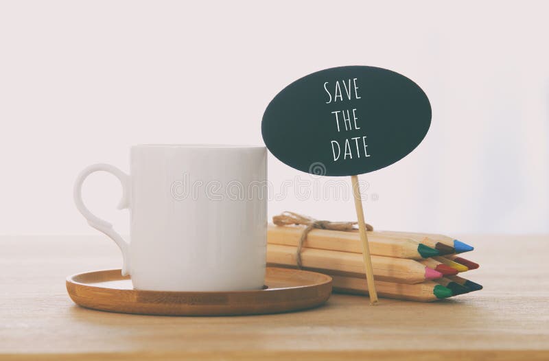 sign with text: SAVE THE DATE next to cup of coffee over wooden table. sign with text: SAVE THE DATE next to cup of coffee over wooden table