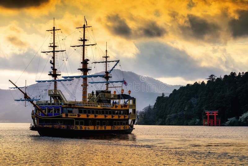 Hakone, Japan - May 4, 2017: Unidentified people on yellow tourist pirate ship floating and sailing on Ashi lake near red torii gate of Hakone shrine with sun rays and twilight sky before sunset. Hakone, Japan - May 4, 2017: Unidentified people on yellow tourist pirate ship floating and sailing on Ashi lake near red torii gate of Hakone shrine with sun rays and twilight sky before sunset.