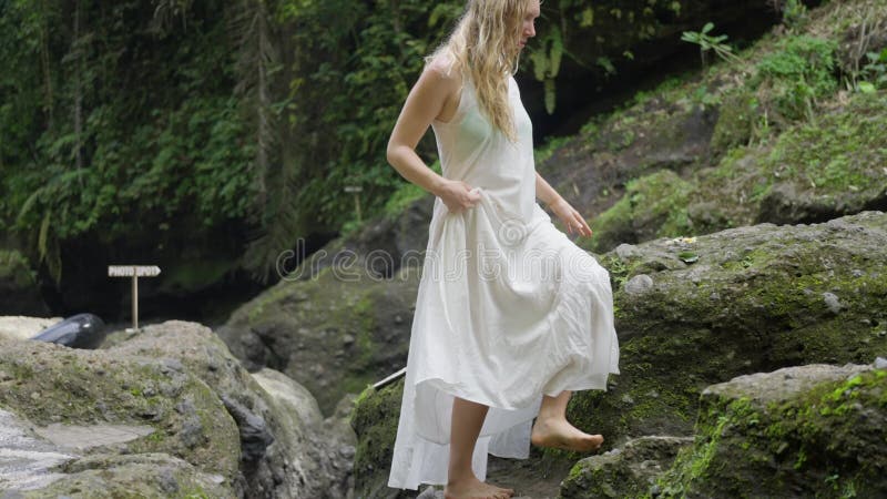 σκανδιναβή γυναίκα με λευκό φόρεμα περπατάει σε βράχους καλυμμένη με πράσινο βρύα