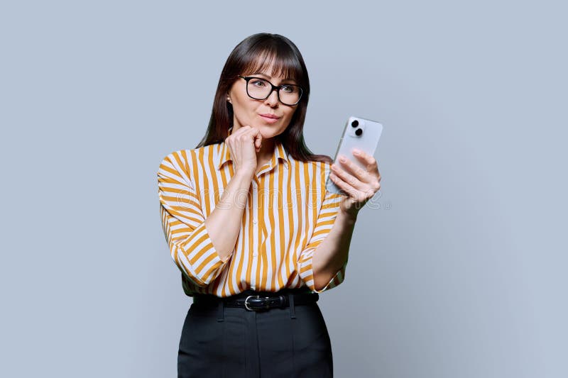 Serious business mature woman using smartphone on gray background. Middle-aged confident successful female holding phone texting reading looking. Mobile internet online technologies apps applications. Serious business mature woman using smartphone on gray background. Middle-aged confident successful female holding phone texting reading looking. Mobile internet online technologies apps applications