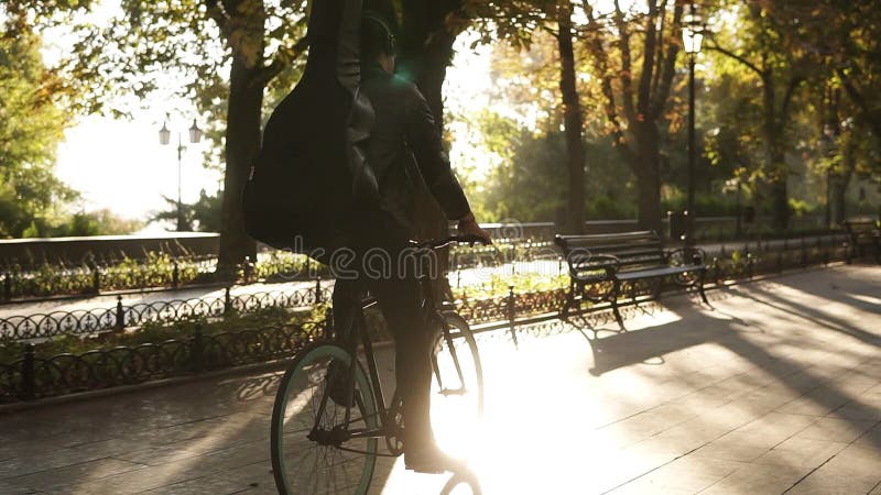 Σπάνια άποψη ενός νεαρού άνδρα στα μαύρα περιστασιακά ενδύματα, ακουστικά που οδηγούν ένα ποδήλατο από το στρωμένο πάρκο πόλεων μ