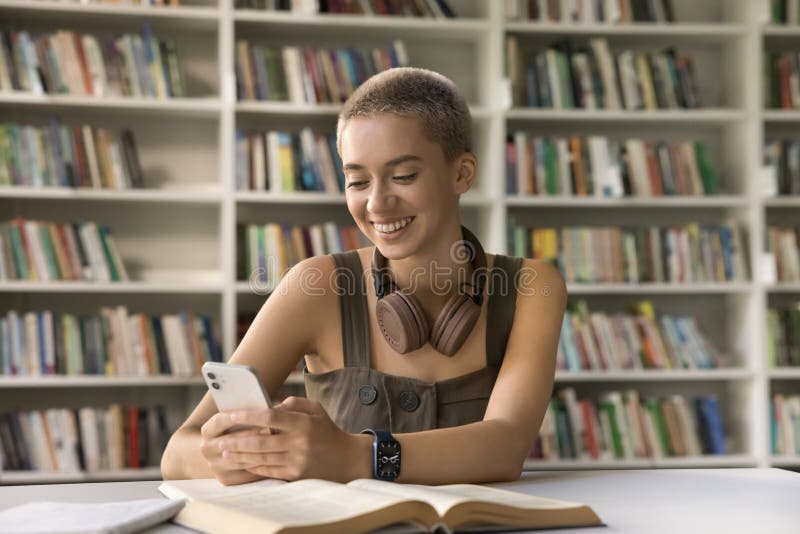 Beautiful short-haired blonde student girl sits at desk holding smart phone, distracted from studying process, chatting on-line with friends, check messages, surfing wireless internet, use mobile apps. Beautiful short-haired blonde student girl sits at desk holding smart phone, distracted from studying process, chatting on-line with friends, check messages, surfing wireless internet, use mobile apps