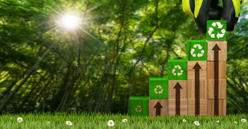 Gloved hand arranging wood blocks with green Recycling Symbols and brown arrows pointing upwards. On a green meadow with a blurry forest in the background. Gloved hand arranging wood blocks with green Recycling Symbols and brown arrows pointing upwards. On a green meadow with a blurry forest in the background