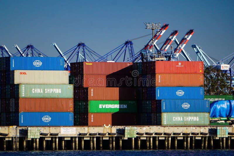 San Pedro, California USA–December 15, 2019: Colorful steel shipping containers sit on a waterside dock at the busy international Port of Los Angeles, industrial equipment and blue sky behind them. San Pedro, California USA–December 15, 2019: Colorful steel shipping containers sit on a waterside dock at the busy international Port of Los Angeles, industrial equipment and blue sky behind them.