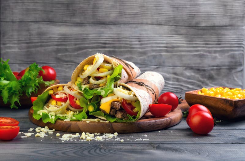 Sandwiches twisted roll Tortilla burritos three pieces on a wooden cutting board on a gray background, lettuce, cucumbers malosollnye, cherry tomatoes, corn, onion rings, meat. Sandwiches twisted roll Tortilla burritos three pieces on a wooden cutting board on a gray background, lettuce, cucumbers malosollnye, cherry tomatoes, corn, onion rings, meat