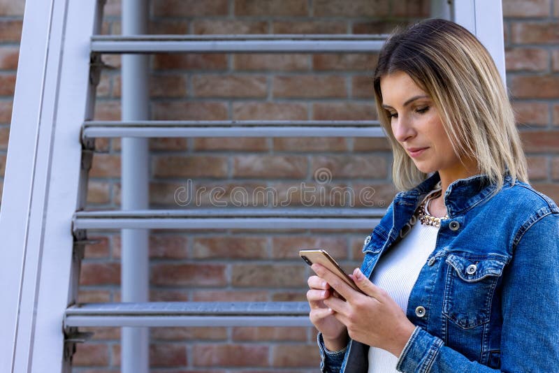 A modern, focused woman interacts with her smartphone, leaning against a metal railing with a brick wall backdrop, illustrating the connectivity of contemporary urban life. Connected World: Modern Woman Using Smartphone in Urban Setting. High quality photo. A modern, focused woman interacts with her smartphone, leaning against a metal railing with a brick wall backdrop, illustrating the connectivity of contemporary urban life. Connected World: Modern Woman Using Smartphone in Urban Setting. High quality photo