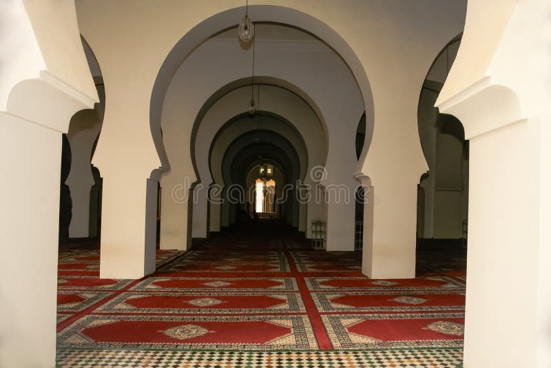 Pale ecru walls make up the continuous archways in succession down the hall, contrasting with continuous and repetitive red rug patterns, inside a Moroccan mosque. Pale ecru walls make up the continuous archways in succession down the hall, contrasting with continuous and repetitive red rug patterns, inside a Moroccan mosque.