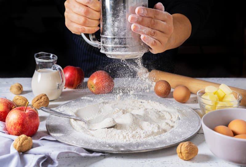 Ingredients for apple cake: apples, eggs, flour, butter, milk and nuts on white wooden background. Woman hands with flour sifter. Ingredients for apple cake: apples, eggs, flour, butter, milk and nuts on white wooden background. Woman hands with flour sifter.