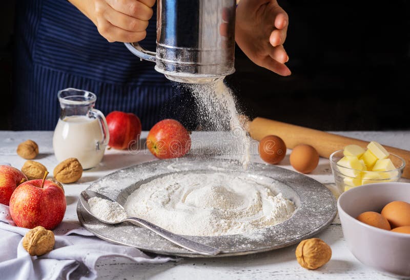 Ingredients for apple cake: apples, eggs, flour, butter, milk and nuts on white wooden background. Woman hands with flour sifter. Ingredients for apple cake: apples, eggs, flour, butter, milk and nuts on white wooden background. Woman hands with flour sifter.