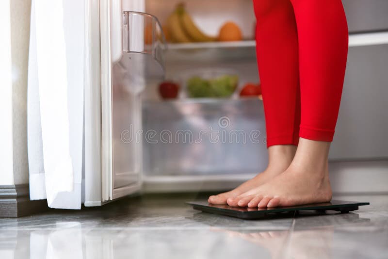 Τα πόδια της γυναίκας στέκονται σε ζυγαριές για να μετρήσουν το βάρος της κουζίνας κοντά σε ανοιχτό ψυγείο γεμάτο φρούτα και λαχα στοκ φωτογραφίες