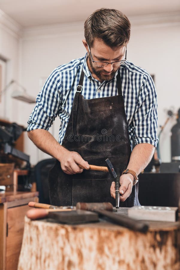 Focused jeweler in an apron using a hammer and a mandrel to shape a new ring while working at a bench in his workshop. Focused jeweler in an apron using a hammer and a mandrel to shape a new ring while working at a bench in his workshop