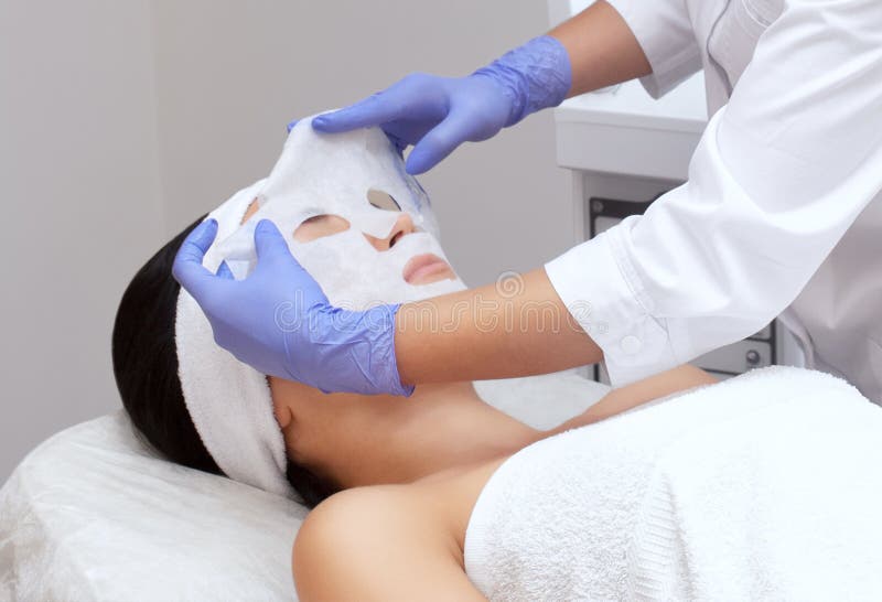 Το cosmetologist για τη διαδικασία και το δέρμα, που εφαρμόζει μια μάσκα φύλλων στο πρόσωπο στοκ εικόνα Το cosmetologist για τη διαδικασία και το δέρμα, που εφαρμόζει μια μάσκα φύλλων στο πρόσωπο στοκ εικόνα