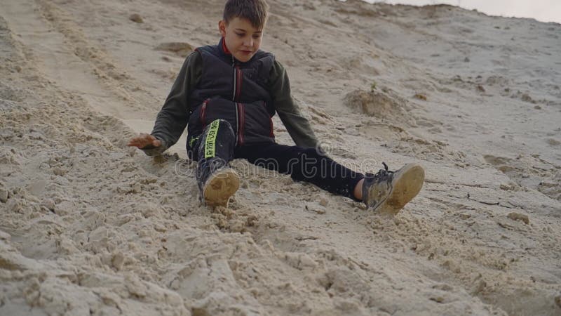 Το αγόρι γλιστράει στην αμμώδη κλίση ενώ κάθεται στην άμμο Εμπρόσθια όψη φιλμ μικρού μήκους