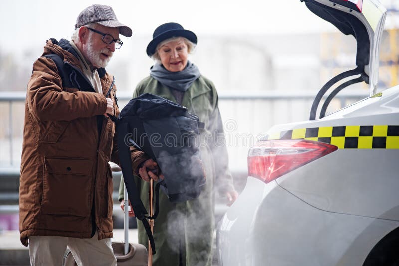 Pleasant gray-haired men in glasses is putting his backpack in trunk of taxi while standing together with his aged wife. Focus on male. Copy space in the right e. Pleasant gray-haired men in glasses is putting his backpack in trunk of taxi while standing together with his aged wife. Focus on male. Copy space in the right e