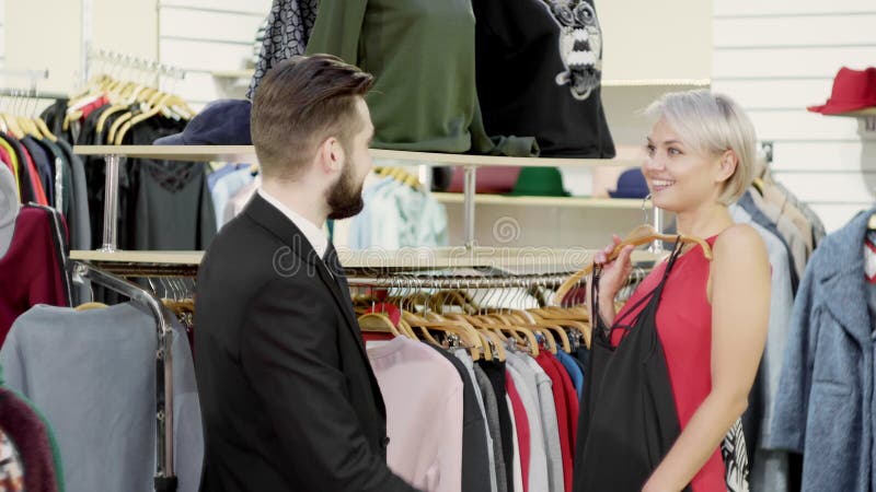 Το ελκυστικό ζεύγος - νέα γυναίκα με το σύζυγό της - επιλέγει τα ενδύματα στο κατάστημα Όμορφη γυναίκα που εξετάζει το φόρεμα απόθεμα βίντεο