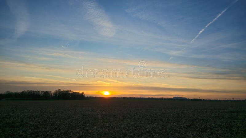 The sun is casting a warm glow as it sets behind a field, with trees silhouetted against the colorful afterglow in the. The sun is casting a warm glow as it sets behind a field, with trees silhouetted against the colorful afterglow in the