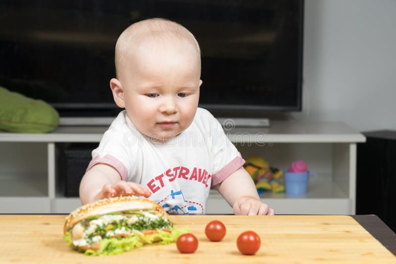 Το καυκάσιο αγόρι νηπίων Litlle προσπαθεί να κλέψει ένα ανθυγειινό εύγευστο χοτ ντογκ με την πράσινη σαλάτα από τον πίνακα στοκ εικόνες