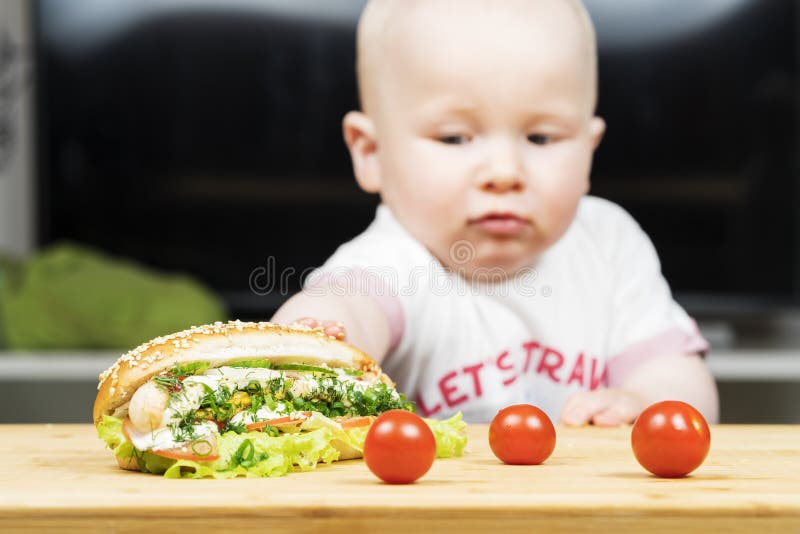 Το καυκάσιο αγόρι νηπίων Litlle προσπαθεί να κλέψει ένα ανθυγειινό εύγευστο χοτ ντογκ με την πράσινη σαλάτα από τον πίνακα στοκ φωτογραφία με δικαίωμα ελεύθερης χρήσης