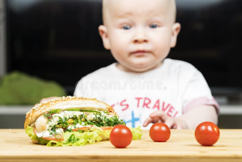 Το καυκάσιο αγόρι νηπίων Litlle προσπαθεί να κλέψει ένα ανθυγειινό εύγευστο χοτ ντογκ με την πράσινη σαλάτα από τον πίνακα στοκ εικόνα