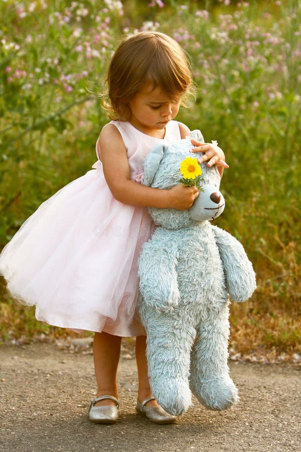 Cute, multi-racial, 2 year old girl wears a dressy, pale pink, full dress and holds a blue stuffed bear and a yellow flower. Little girl is Asian/Caucasian with brown hair and eyes. Green plants and flowers background. Cute, multi-racial, 2 year old girl wears a dressy, pale pink, full dress and holds a blue stuffed bear and a yellow flower. Little girl is Asian/Caucasian with brown hair and eyes. Green plants and flowers background.