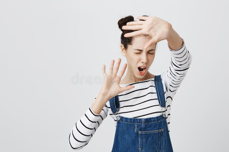 Displeased brunette girl with hairbuns wearing stylish denim clothes posing against gray studio wall, frowning face in displeasure, keeping hands in stop gesture, as if saying: Stay away from me. Body language. Displeased brunette girl with hairbuns wearing stylish denim clothes posing against gray studio wall, frowning face in displeasure, keeping hands in stop gesture, as if saying: Stay away from me. Body language.