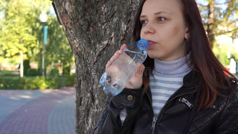 Το κορίτσι Brunette πίνει το πόσιμο νερό από το μπουκάλι στο πράσινο θερινό πάρκο Υγιής τρόπος ζωής Έννοια καθαρού νερού κατανάλω