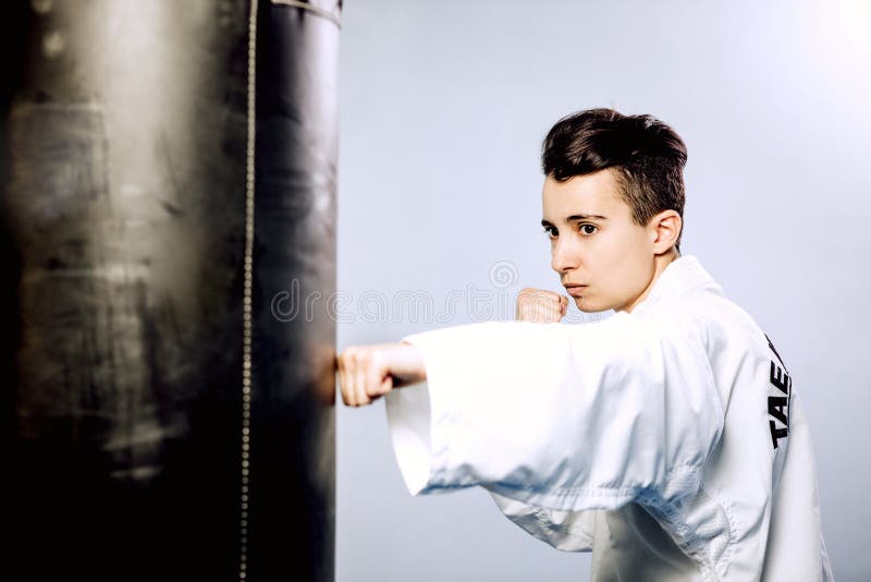 Το κορίτσι σε ένα άσπρο κιμονό κτυπά τη μαύρη punching τσάντα στο γκρίζο υπόβαθρο Από Tae Kwon, karate, κλωτσά στοκ εικόνες με δικαίωμα ελεύθερης χρήσης Το κορίτσι σε ένα άσπρο κιμονό κτυπά τη μαύρη punching τσάντα στο γκρίζο υπόβαθρο Από Tae Kwon, karate, κλωτσά στοκ εικόνες με δικαίωμα ελεύθερης χρήσης