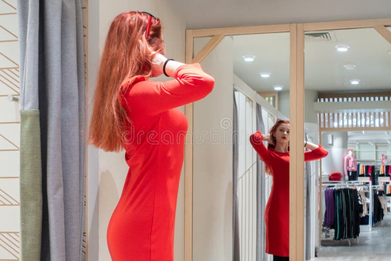 Το λεπτό κορίτσι προσπαθεί σε ένα φόρεμα στο κατάστημα η κοκκινομάλλης γυναίκα σε ένα κόκκινο φόρεμα θέτει κοντά σε έναν καθρέφτη στοκ φωτογραφίες με δικαίωμα ελεύθερης χρήσης Το λεπτό κορίτσι προσπαθεί σε ένα φόρεμα στο κατάστημα η κοκκινομάλλης γυναίκα σε ένα κόκκινο φόρεμα θέτει κοντά σε έναν καθρέφτη στοκ φωτογραφίες με δικαίωμα ελεύθερης χρήσης
