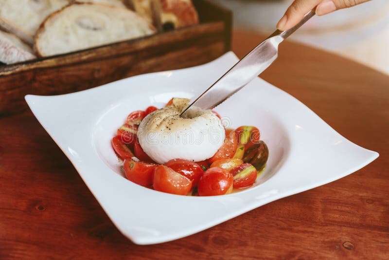 Το μαχαίρι που κόβει το τυρί Burrata με τη σαλάτα ντοματών κερασιών confit εξυπηρέτησε στο άσπρο πιάτο στον ξύλινο πίνακα στοκ εικόνα Το μαχαίρι που κόβει το τυρί Burrata με τη σαλάτα ντοματών κερασιών confit εξυπηρέτησε στο άσπρο πιάτο στον ξύλινο πίνακα στοκ εικόνα