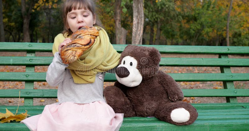 Το μικρό κορίτσι τρώει το πλεγμένο κουλούρι με την παπαρούνα στον πάγκο με τη teddy αρκούδα στην παιδική χαρά στο πάρκο φθινοπώρο