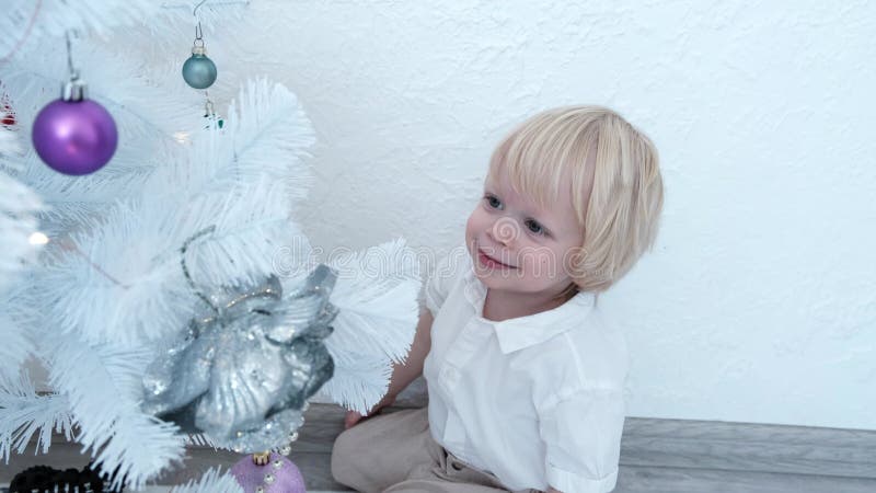 το μωρό κάθεται κοντά σε ένα λευκό χριστουγεννιάτικο δέντρο και παίζει μαζί του