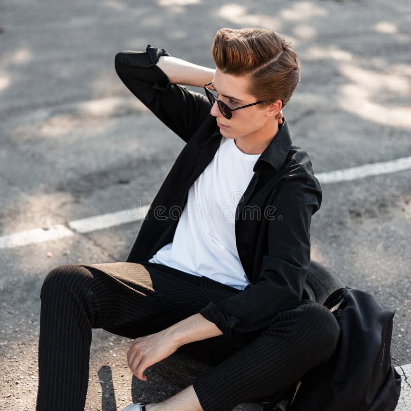 Το νέο άτομο hipster στο καθιερώνον τη μόδα πουκάμισο στο παντελόνι με το σακίδιο πλάτης με το hairstyle θέτει τη συνεδρίαση στην στοκ φωτογραφία με δικαίωμα ελεύθερης χρήσης Το νέο άτομο hipster στο καθιερώνον τη μόδα πουκάμισο στο παντελόνι με το σακίδιο πλάτης με το hairstyle θέτει τη συνεδρίαση στην στοκ φωτογραφία με δικαίωμα ελεύθερης χρήσης
