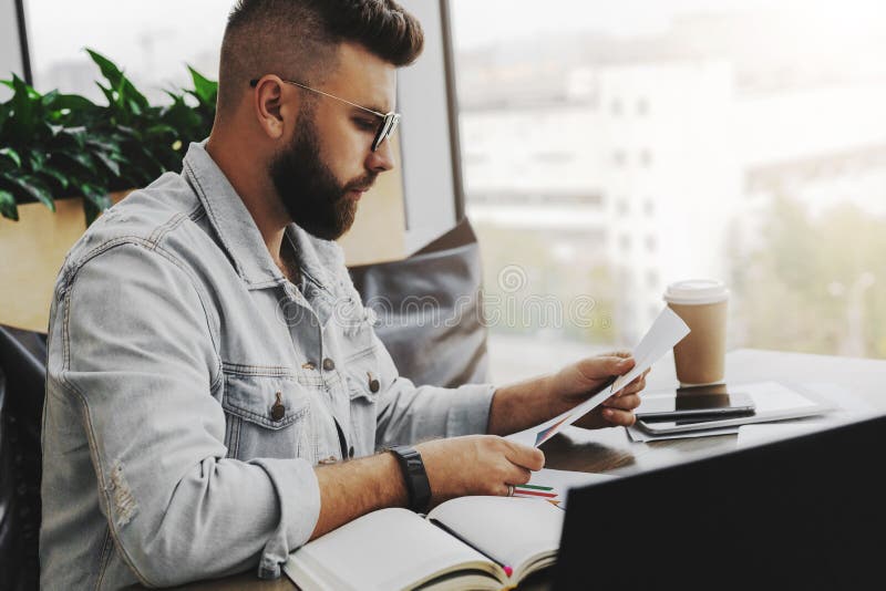Το νέο ελκυστικό γενειοφόρο άτομο hipster κάθεται στον καφέ μπροστά από το aptop, διαβάζοντας τα έγγραφα εγγράφου Το Freelancer λ στοκ φωτογραφίες με δικαίωμα ελεύθερης χρήσης Το νέο ελκυστικό γενειοφόρο άτομο hipster κάθεται στον καφέ μπροστά από το aptop, διαβάζοντας τα έγγραφα εγγράφου Το Freelancer λ στοκ φωτογραφίες με δικαίωμα ελεύθερης χρήσης