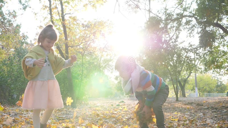 Το πάρκο πόλεων φθινοπώρου, παιδιά πηδά και ρίχνει τα φύλλα στο υπόβαθρο των δέντρων και του φυλλώματος στον ήλιο