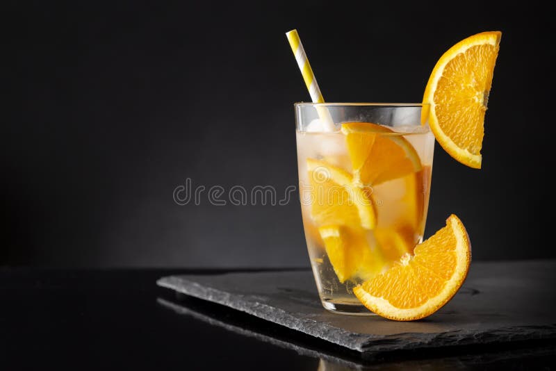 Glass of fresh, cold infused water with orange slices, ice cubes and a drinking straw on a black stone tray. Glass of fresh, cold infused water with orange slices, ice cubes and a drinking straw on a black stone tray