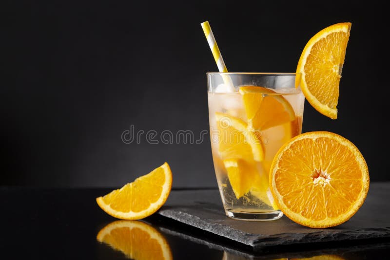 Glass of fresh, cold infused water with orange slices, ice cubes and a drinking straw on a black stone tray. Glass of fresh, cold infused water with orange slices, ice cubes and a drinking straw on a black stone tray