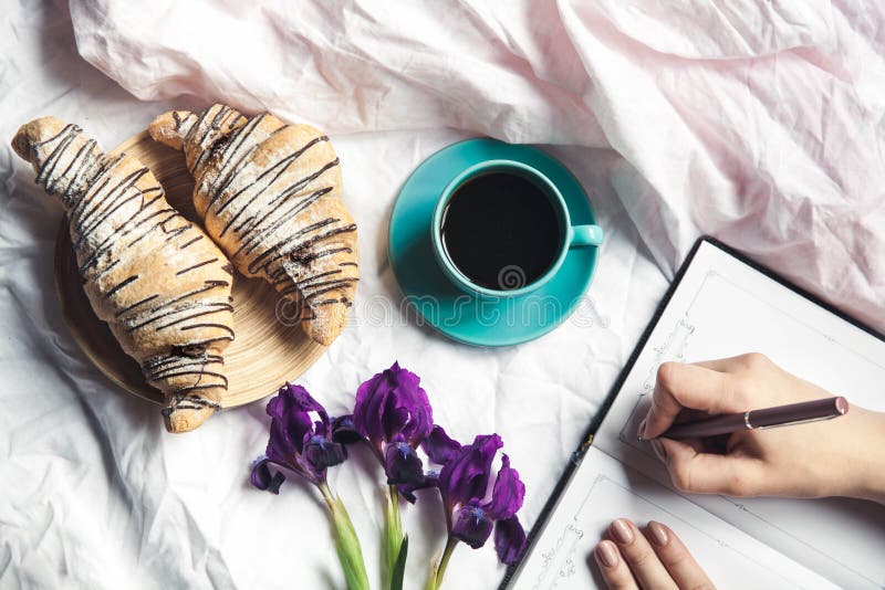 Το πρόγευμα στο κρεβάτι, ένας croissant με τον καφέ και τα λουλούδια στο κρεβάτι, παίρνουν τις σημειώσεις σε ένα σημειωματάριο στοκ εικόνες Το πρόγευμα στο κρεβάτι, ένας croissant με τον καφέ και τα λουλούδια στο κρεβάτι, παίρνουν τις σημειώσεις σε ένα σημειωματάριο στοκ εικόνες