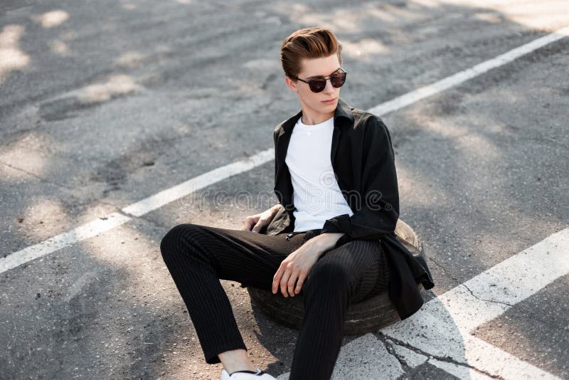 Το σύγχρονο νέο άτομο hipster στα γυαλιά ηλίου στα μοντέρνα ενδύματα με ένα καθιερώνον τη μόδα hairstyle χαλαρώνει τη συνεδρίαση στοκ εικόνες Το σύγχρονο νέο άτομο hipster στα γυαλιά ηλίου στα μοντέρνα ενδύματα με ένα καθιερώνον τη μόδα hairstyle χαλαρώνει τη συνεδρίαση στοκ εικόνες